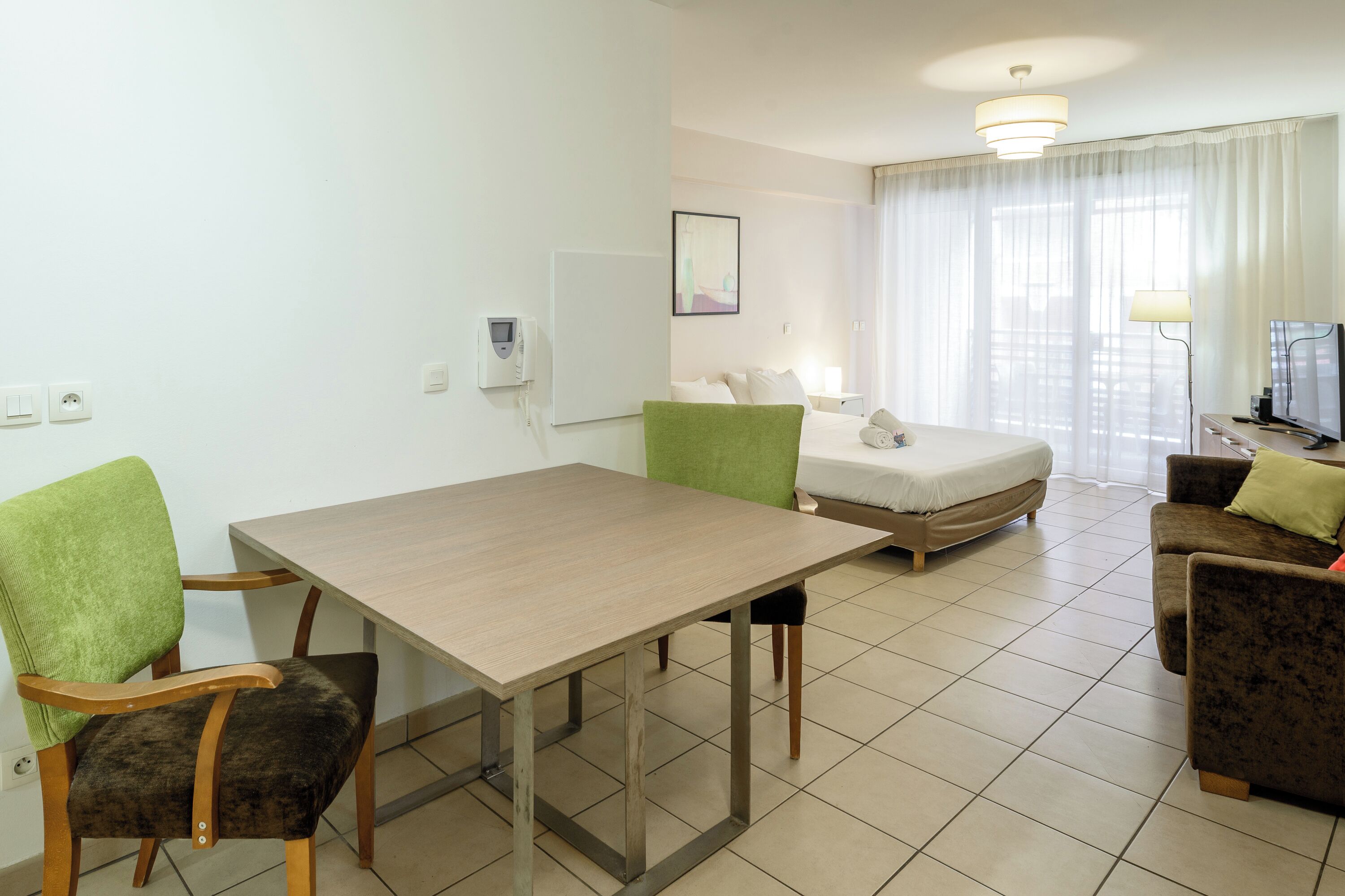 A modern hotel room with a dining table, two chairs, a sofa, a bed, and a television.