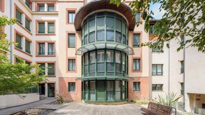 Un edificio de varios pisos con un amplio ventanal curvo, un patio con bancos y fachada de ladrillo.