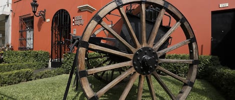 A large wooden wheel with a metal hub, positioned in front of a building with a red facade, black iron gates, and a sign that reads "Casa de la Cultura."