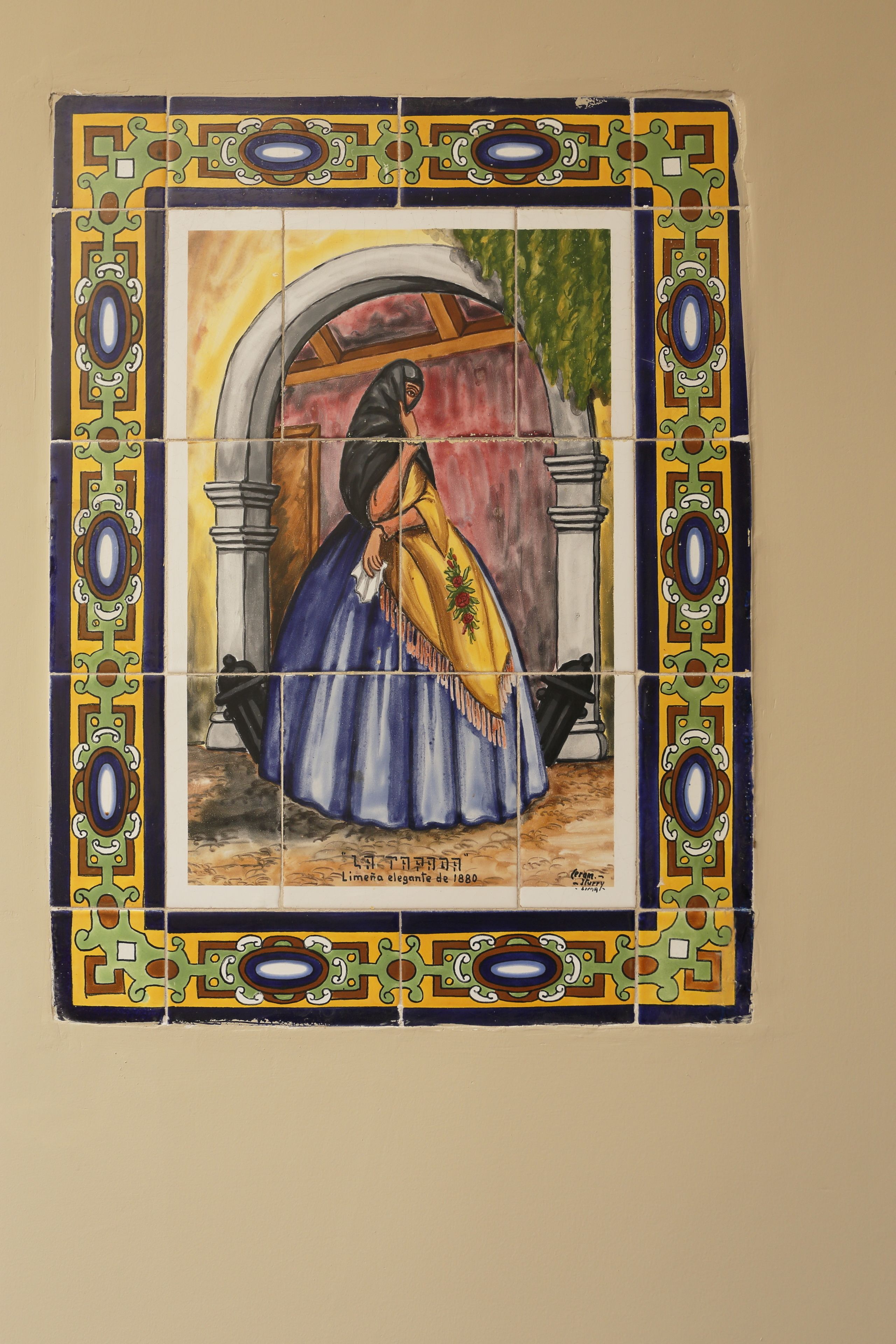 Eine mit Fliesen verkleidete Wand mit dekorativer Bordüre und einem zentralen Feld, das eine Frau in einem blauen Kleid zeigt.