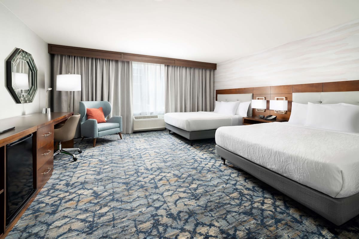 Een hotelkamer met een groot bed, een kleiner bed, een bureau met stoel, een televisie en een raam met gordijnen.