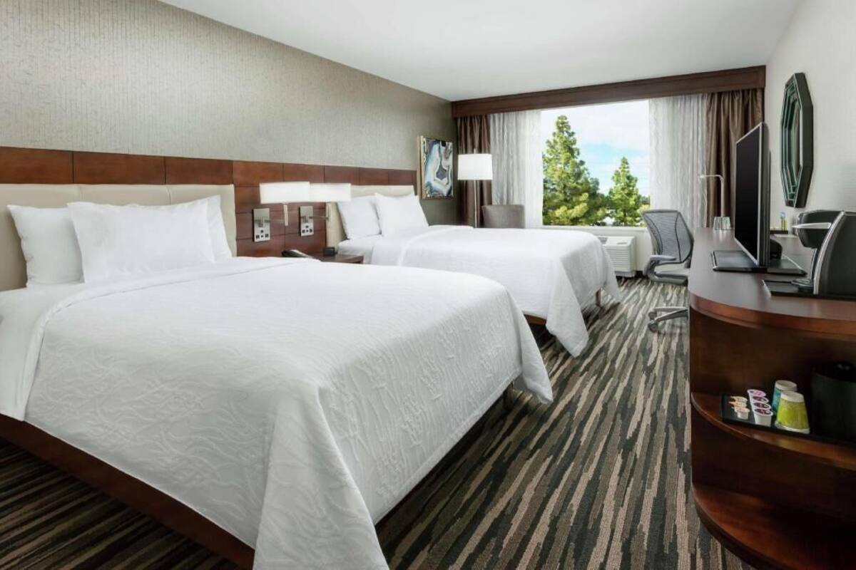 Hotelkamer met twee bedden, een bureau met stoel, een televisie en een raam met uitzicht op bomen.