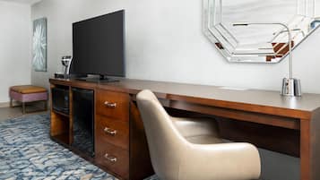 A modern hotel room with a wooden desk, a flat-screen TV, a built-in mirror, and a leather chair.