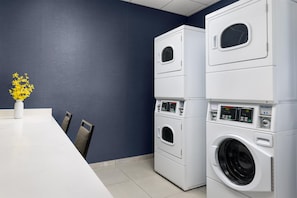A laundry room with stacked washing machines and dryers, a white table with chairs, and a vase with yellow flowers.