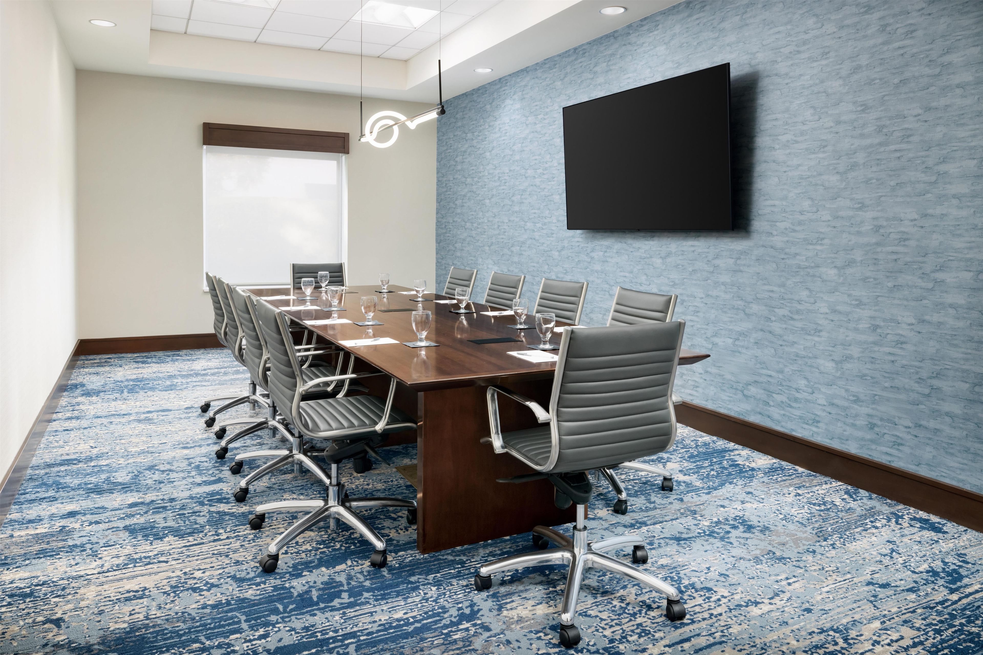 A conference room with a long table, multiple chairs, a flat-screen TV mounted on the wall, and a patterned carpet.