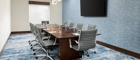 A conference room with a long table, multiple chairs, a flat-screen TV mounted on the wall, and a patterned carpet.