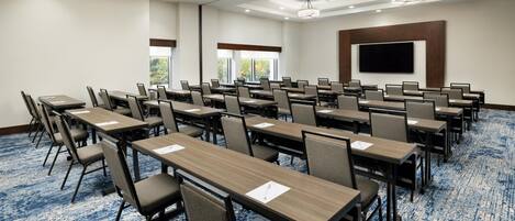 Un salón de conferencias con filas de mesas y sillas, una pantalla grande y piso alfombrado.