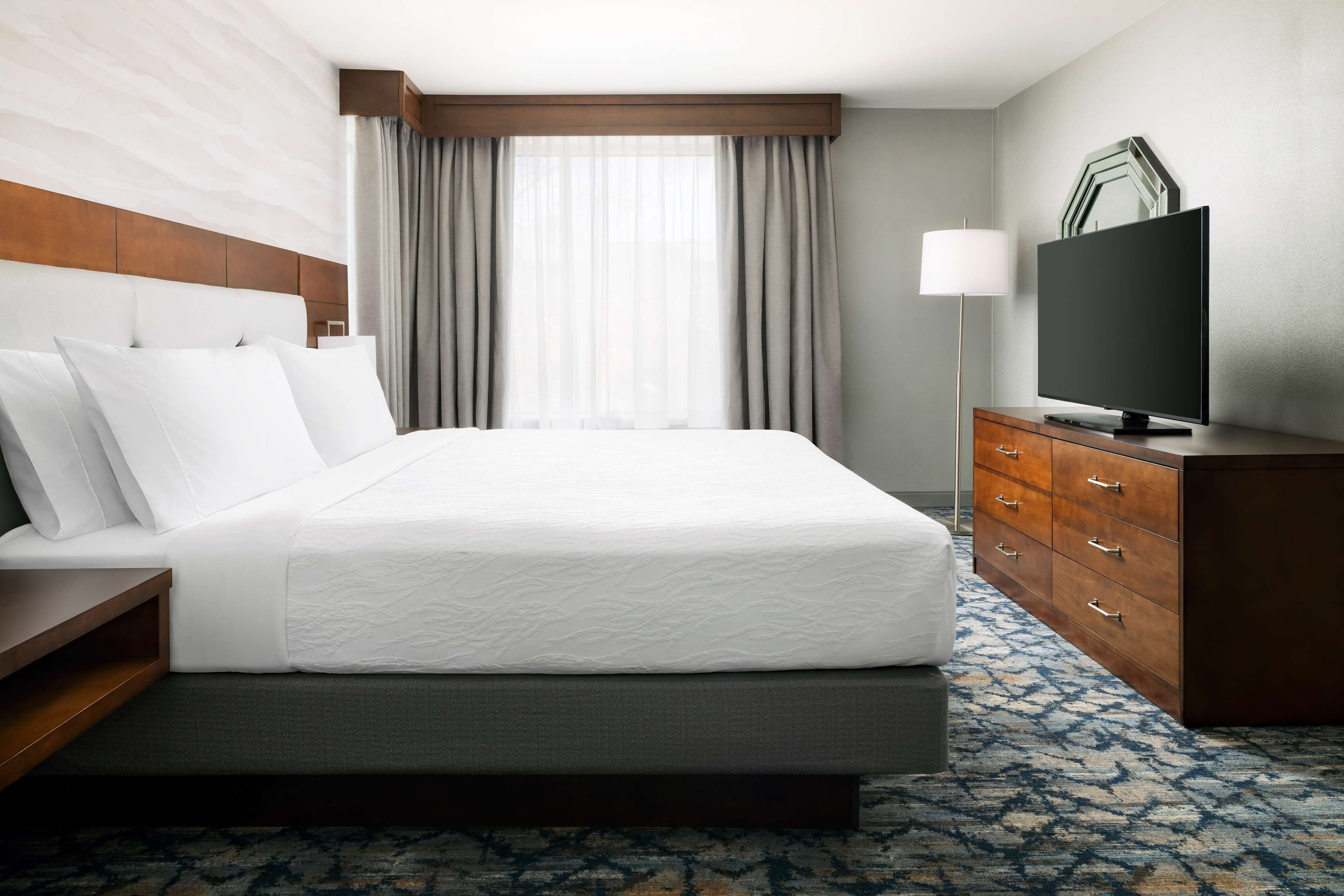 A hotel room with a large bed, a wooden headboard, a flat-screen TV, a desk, and a dresser.
