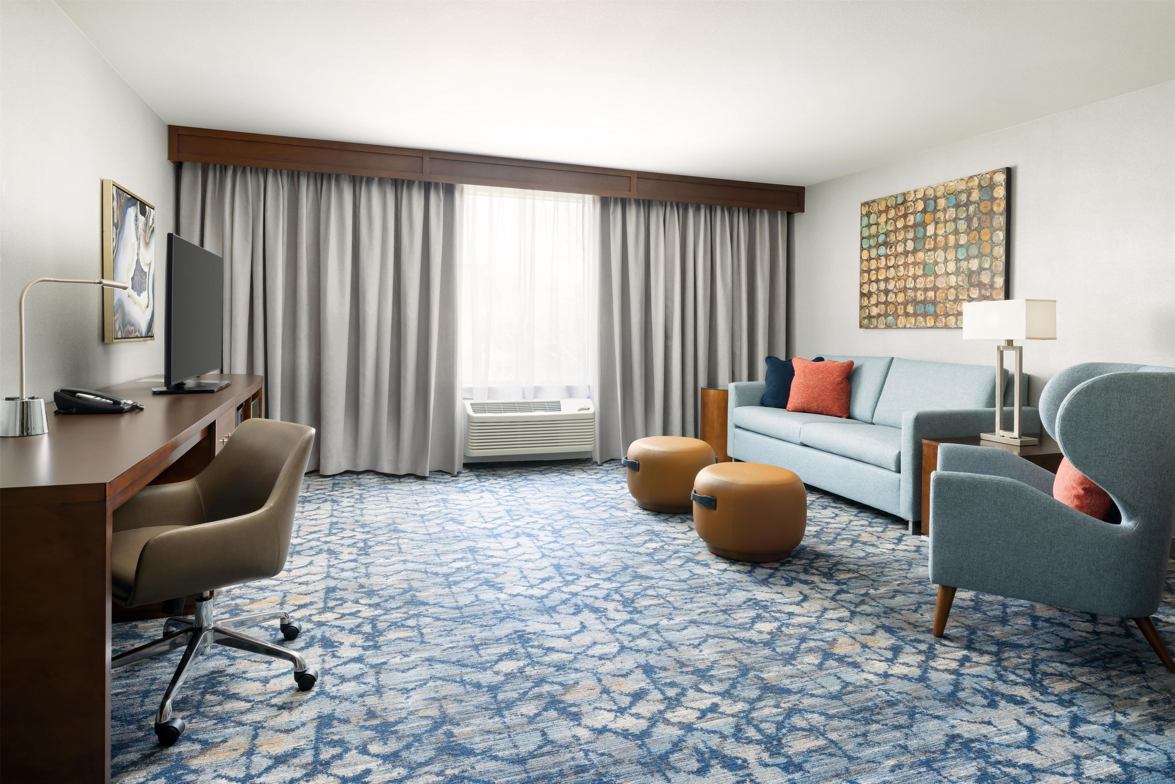 Una habitación de hotel con escritorio, sofá, dos otomanas, televisor y un ventanal con cortinas.