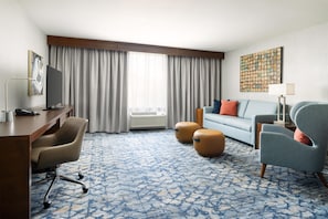 A hotel room with a desk, a sofa, two ottomans, a TV, and a large window with curtains. - Hilton Garden Inn Palo Alto (Palo Alto)
