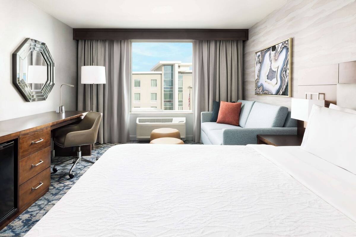 Een hotelkamer met een groot bed, een bureau met lades, een stoel, een klein voetenbankje, een bank met kussens, een groot raam met gordijnen en een wandlamp.