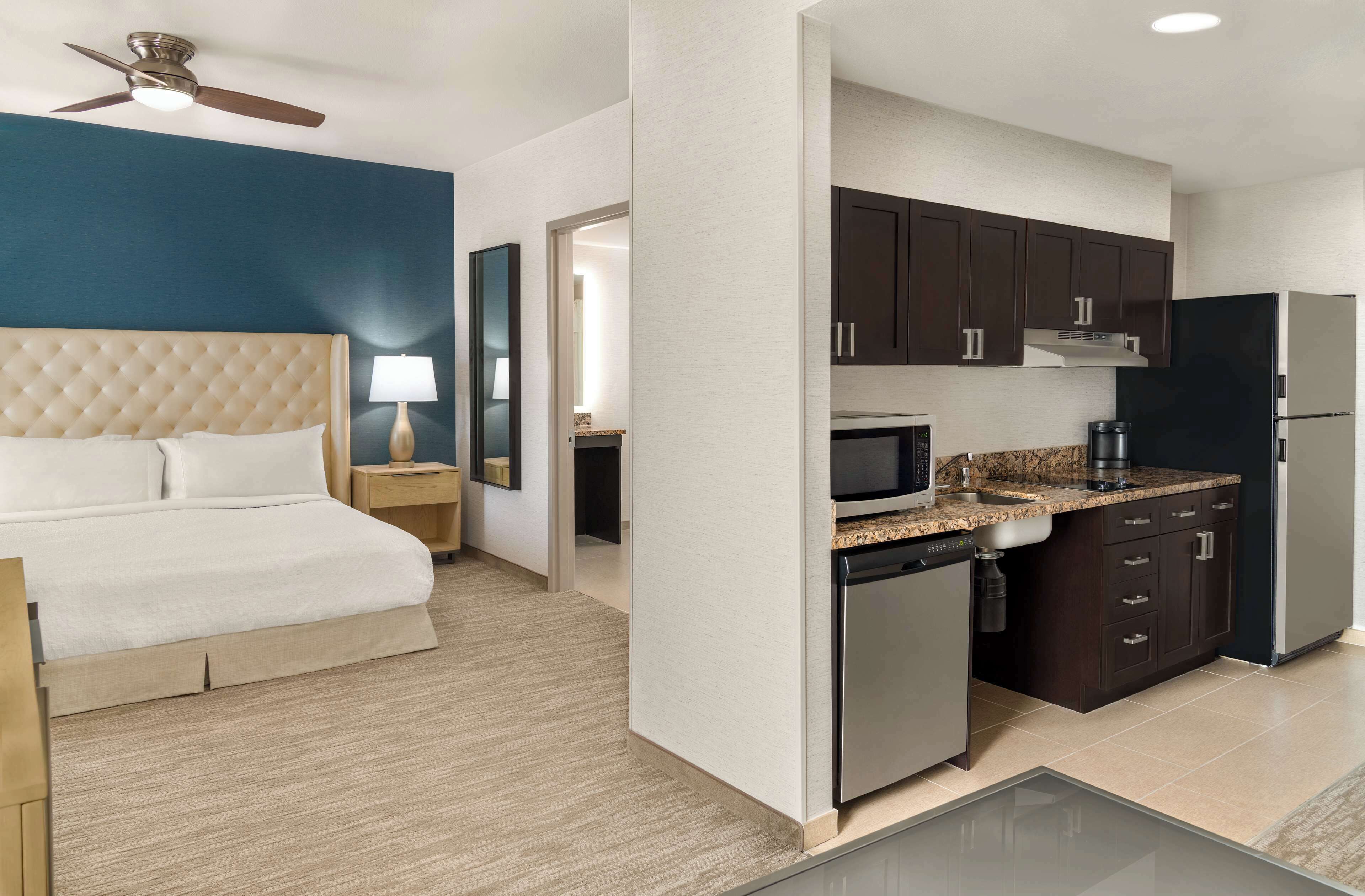 A hotel room with a bed, a nightstand, a lamp, a kitchenette with a microwave, stove, and refrigerator.