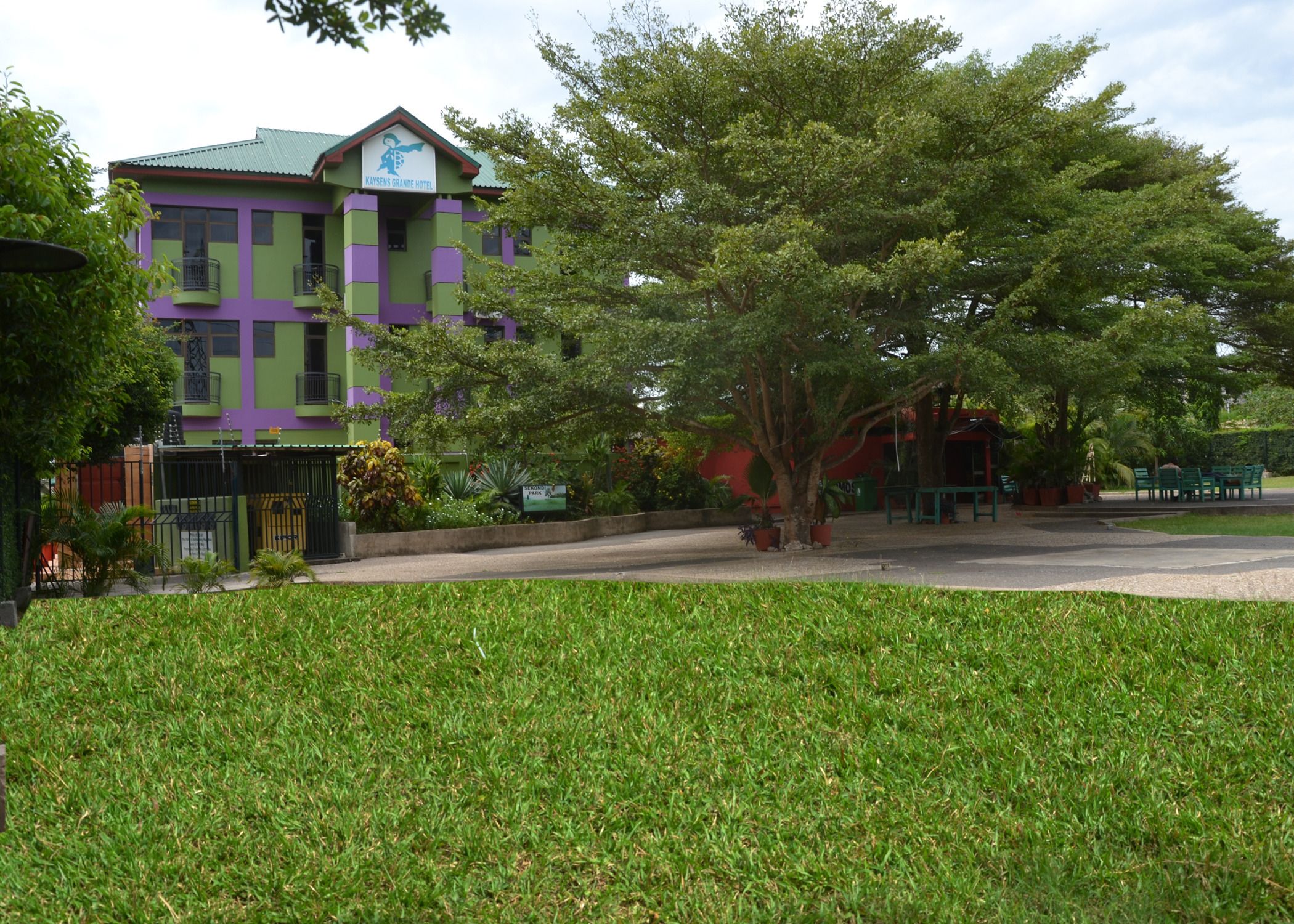 Un hotel con fachada verde y morada, césped cuidado y mobiliario exterior.