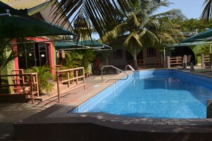 Una piscina con borde de azulejos, rodeada de palmeras y zonas de estar cubiertas.