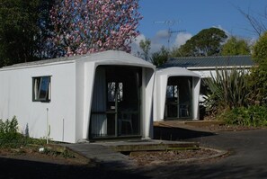 A white mobile home with a large window and a door, surrounded by vegetation and a tree with pink flowers. - Cottage Park Motor Lodge and Conference Centre (Otaki)