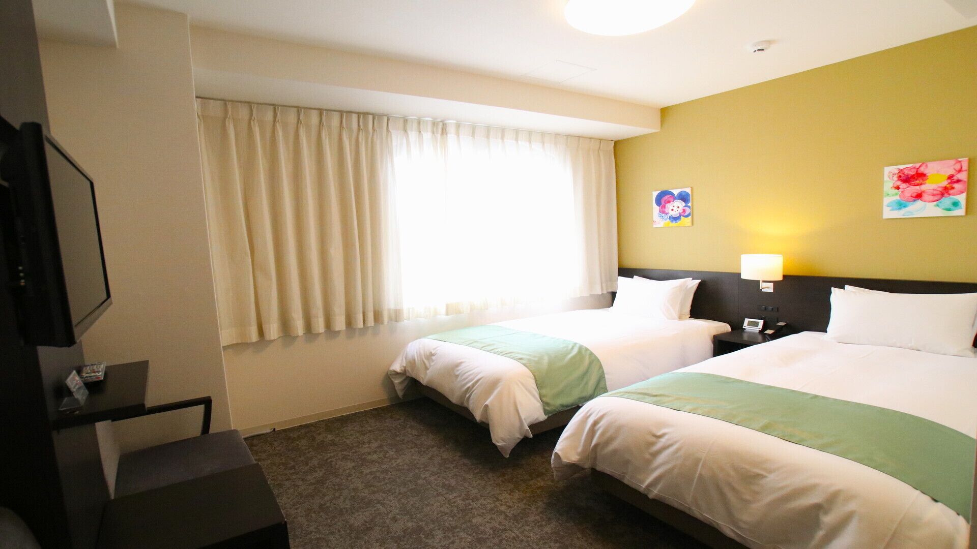 Ein Hotelzimmer mit zwei Betten, einem Fernseher, einem Schreibtisch, einem Fenster mit Vorhängen und zwei Wandbildern.