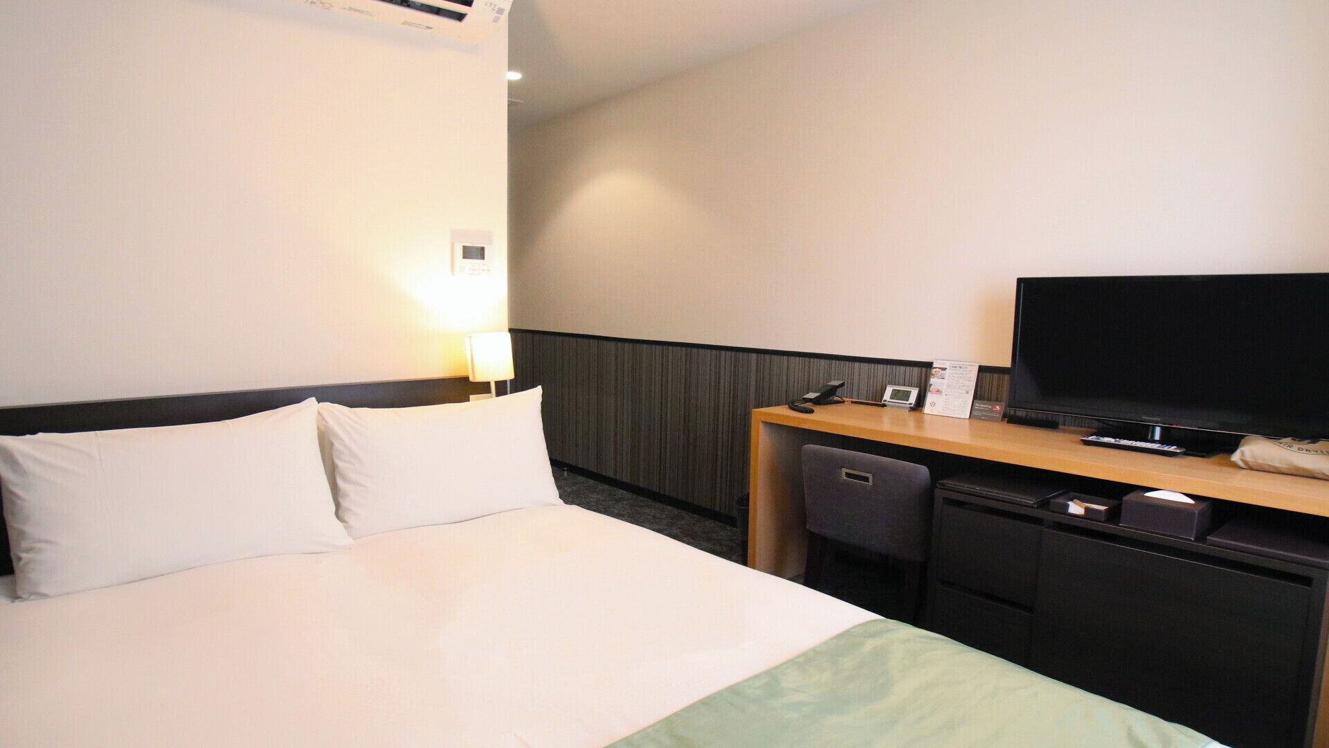 A hotel room with a bed, a desk with a TV, a chair, and a small desk lamp.