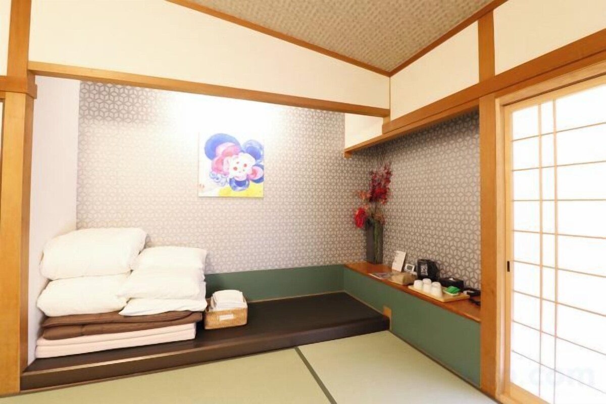 A traditional Japanese room with tatami flooring, a low bed, a small table with a vase, and a sliding door.