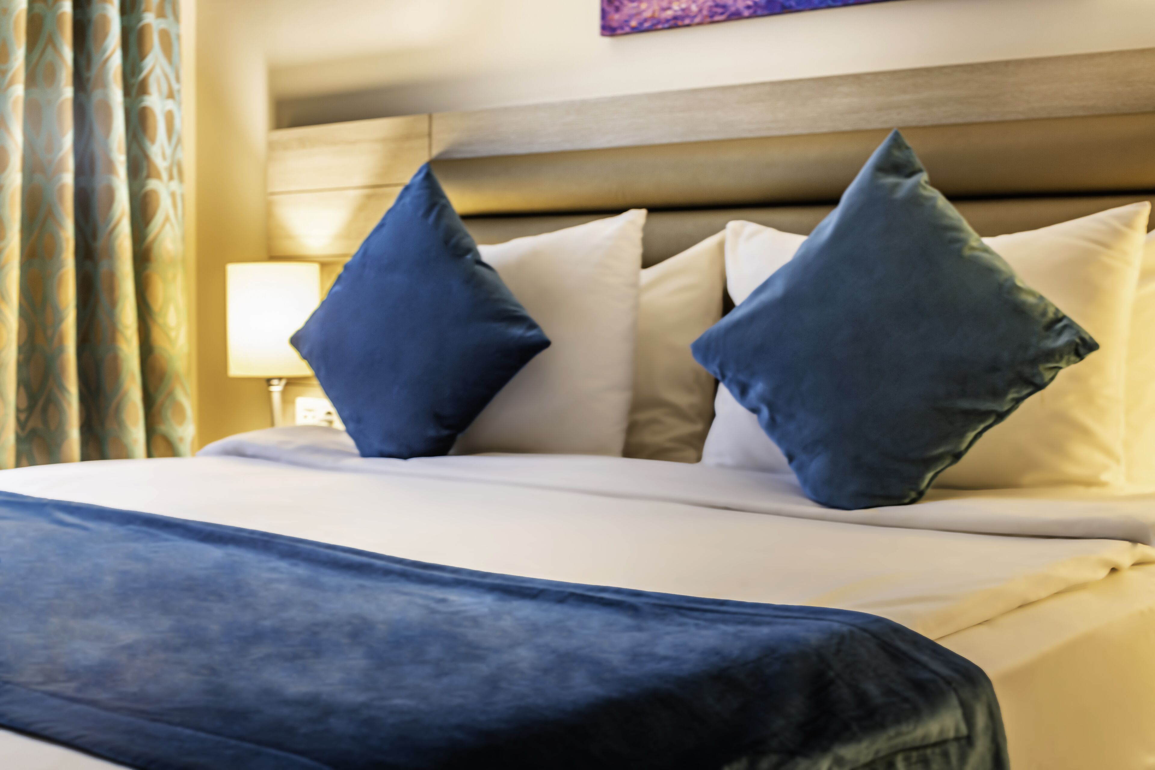 A neatly made bed with a blue bedspread and two pillows, a wooden headboard, and a lamp on a bedside table.