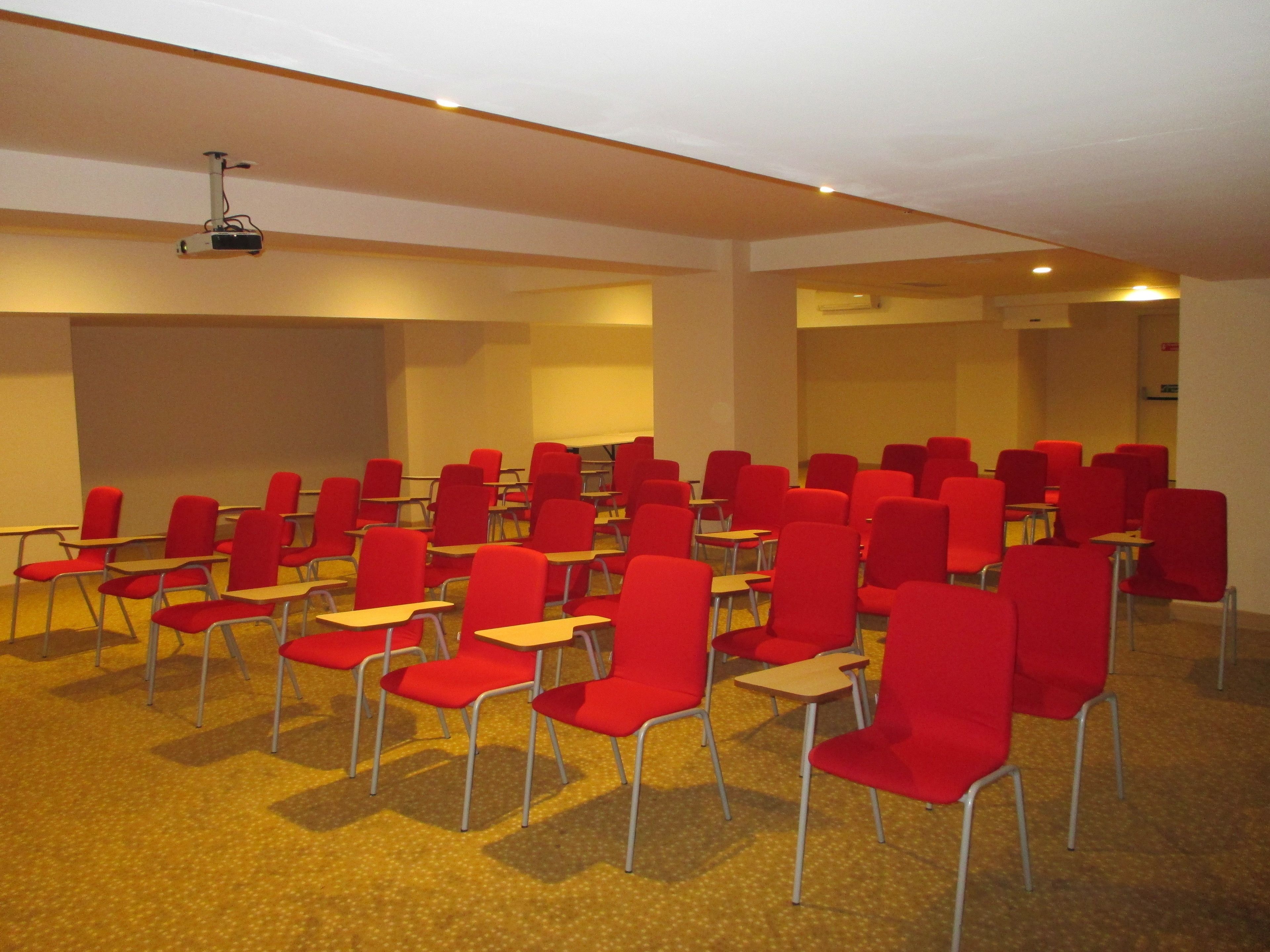 A conference room with rows of red chairs and tables, a projector mounted on the ceiling, and carpeted flooring.