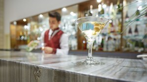 A martini glass on a marble countertop in a bar area with a blurred background of bottles and a person.