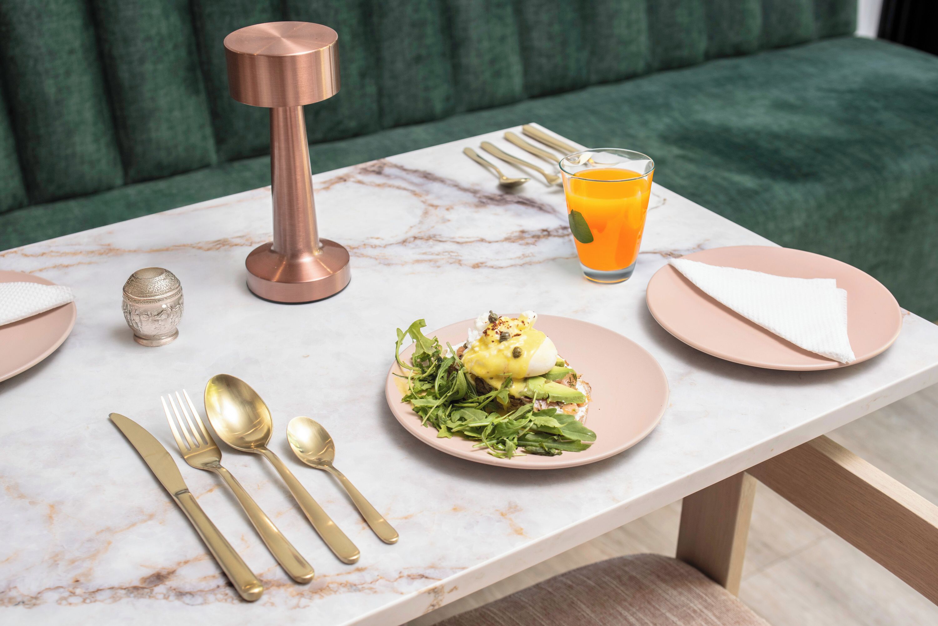 A dining table set with gold cutlery, a pink plate with food, a glass of orange juice, and a copper candle holder.