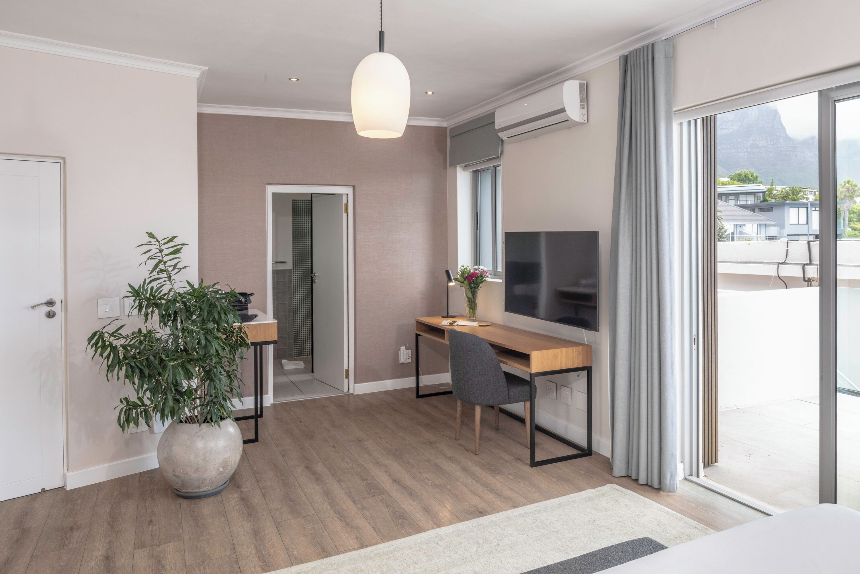 Une chambre d’hôtel moderne avec un bureau en bois, une grande plante en pot, un téléviseur à écran plat et une porte-fenêtre coulissante donnant sur un balcon extérieur.