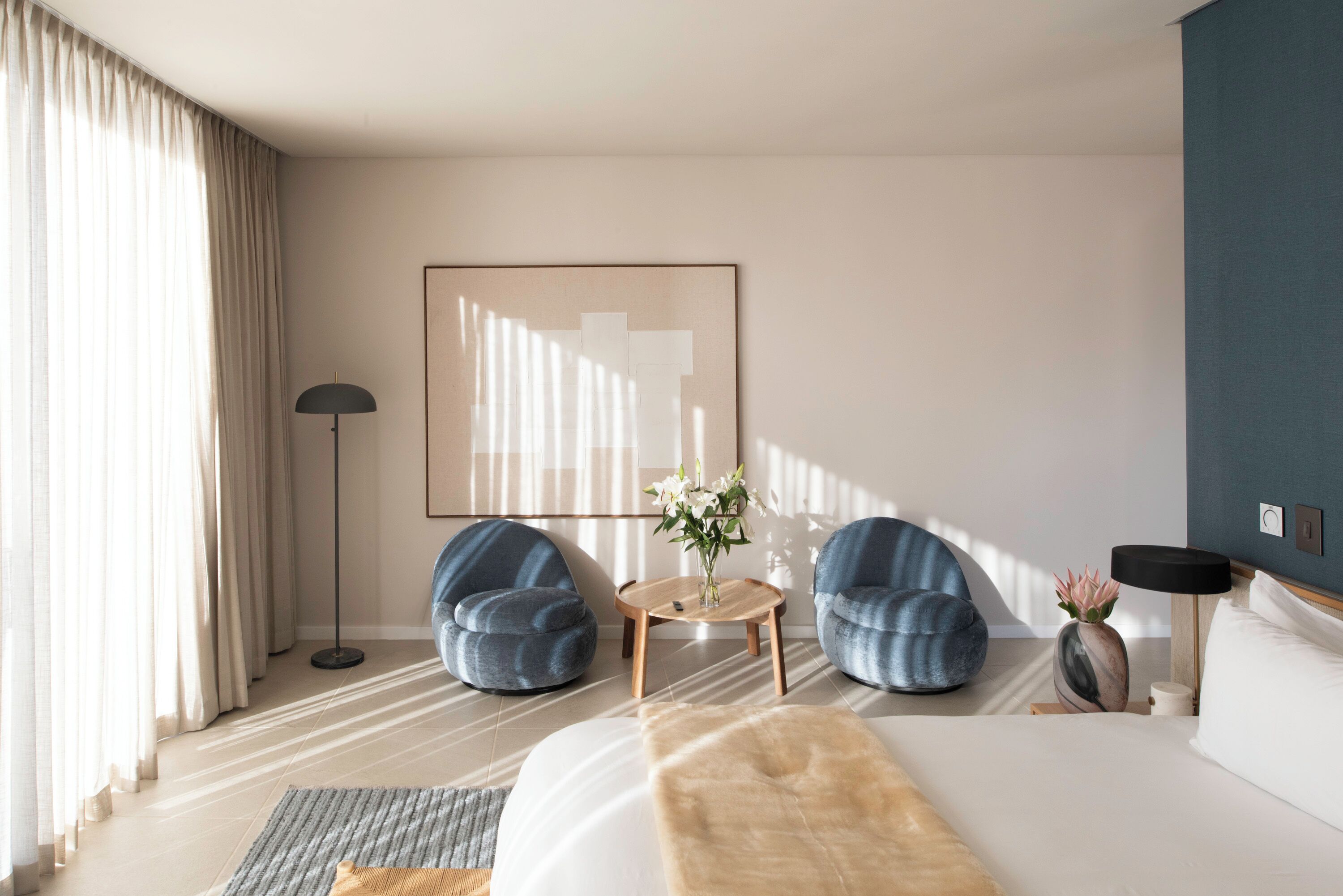 A modern hotel room with a large bed, two blue armchairs, a small round table, and a floor lamp.