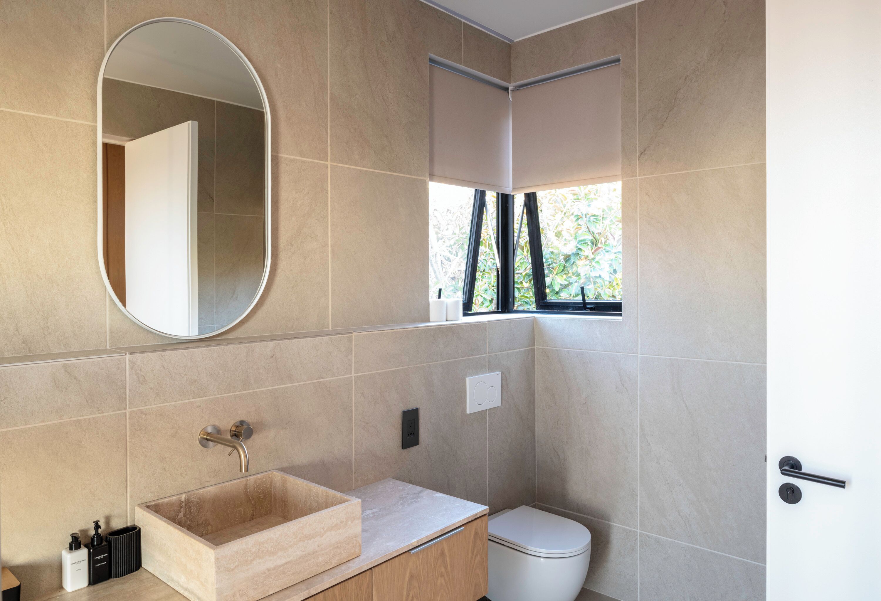 Une salle de bain moderne avec un mur recouvert de carreaux beiges, un lavabo rectangulaire avec un meuble-lavabo en bois, des toilettes blanches et une fenêtre donnant sur un paysage verdoyant.