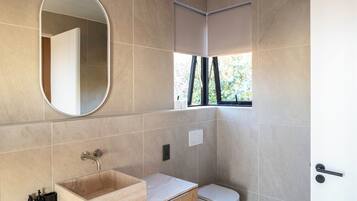 Een moderne badkamer met een behangkleurige betegelde muur, een rechthoekig wastafelmeubel van hout, een wit toilet en een raam met uitzicht op groen.