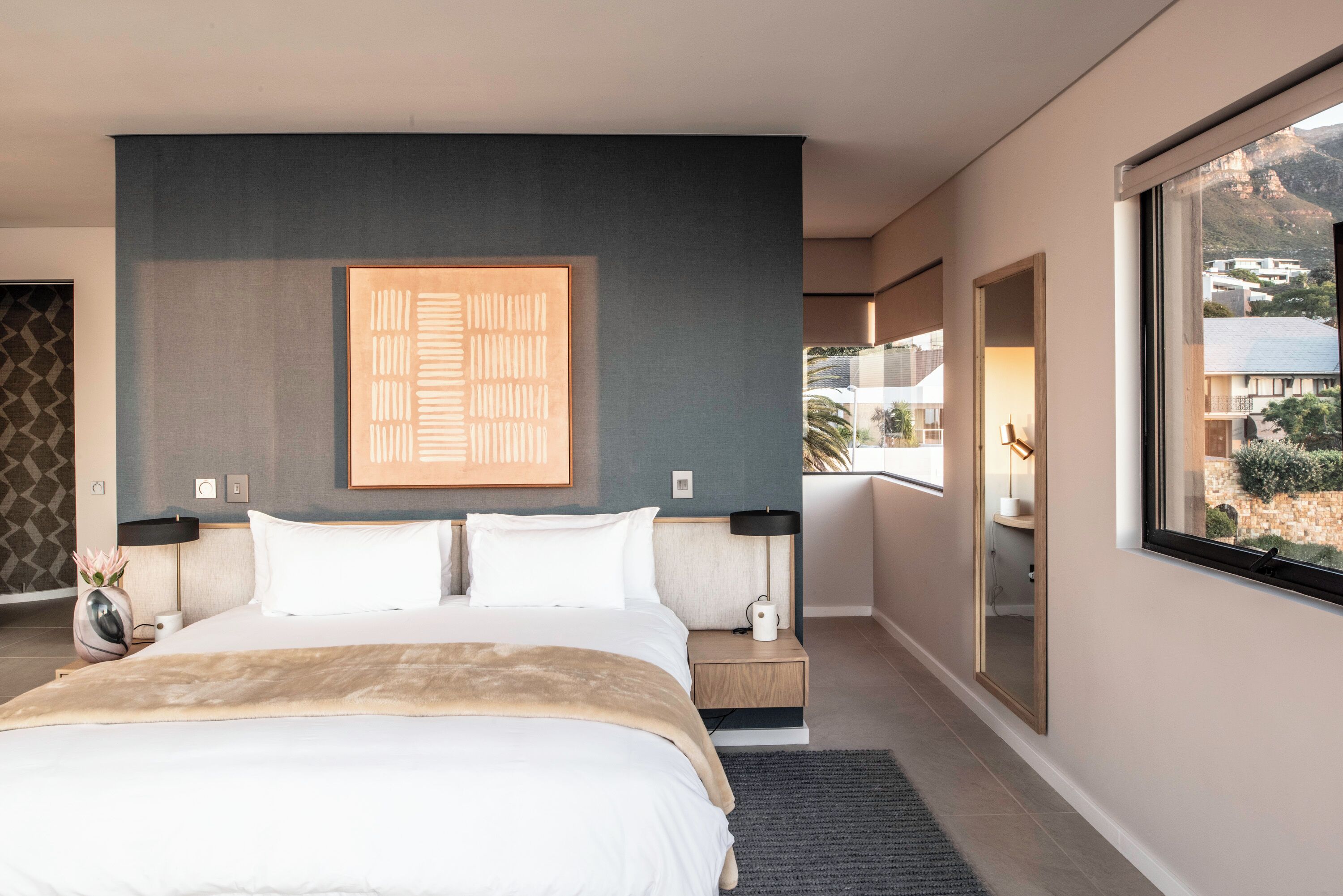 A modern hotel room with a large bed, a nightstand, a bedside table, and a large window offering a view of the city.