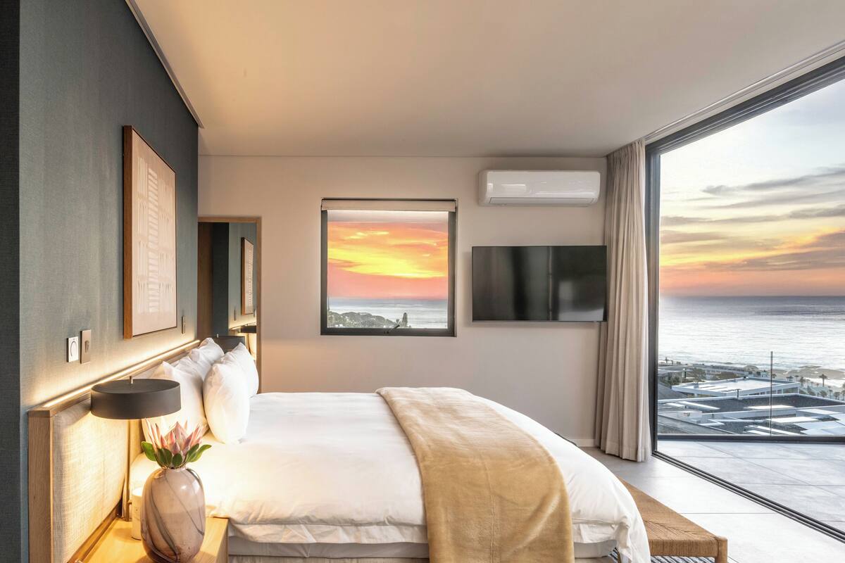 Moderni hotellihuone, jossa on suuri sänky, yöpöytä lampun kanssa, maljakko kukkineen ja suuri ikkuna, josta avautuu näkymä auringonlaskuun.