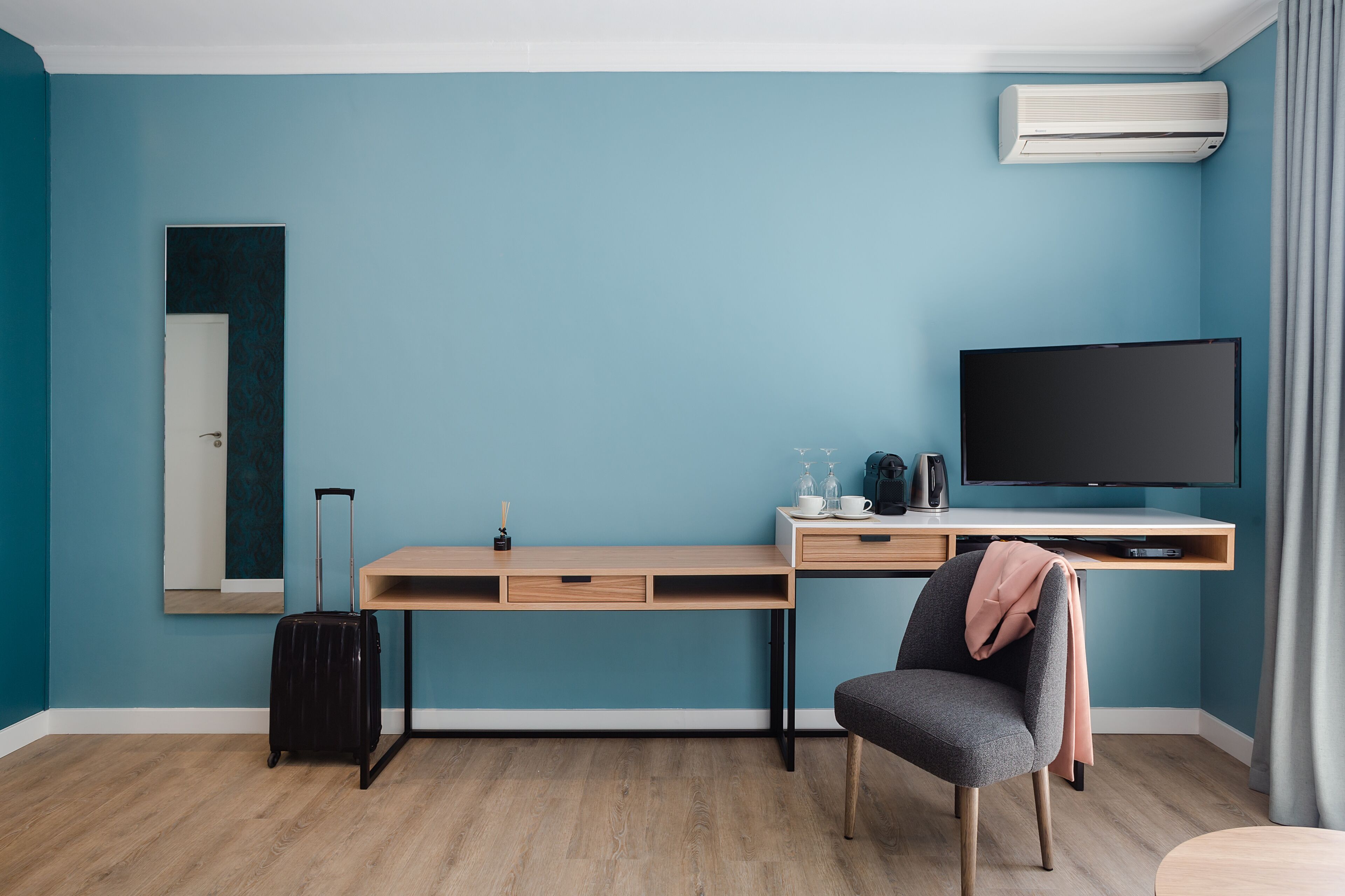 Een moderne hotelkamer met een blauwgroene wand, een houten bureau, een televisie, een stoel, een koffer en een airconditioner.