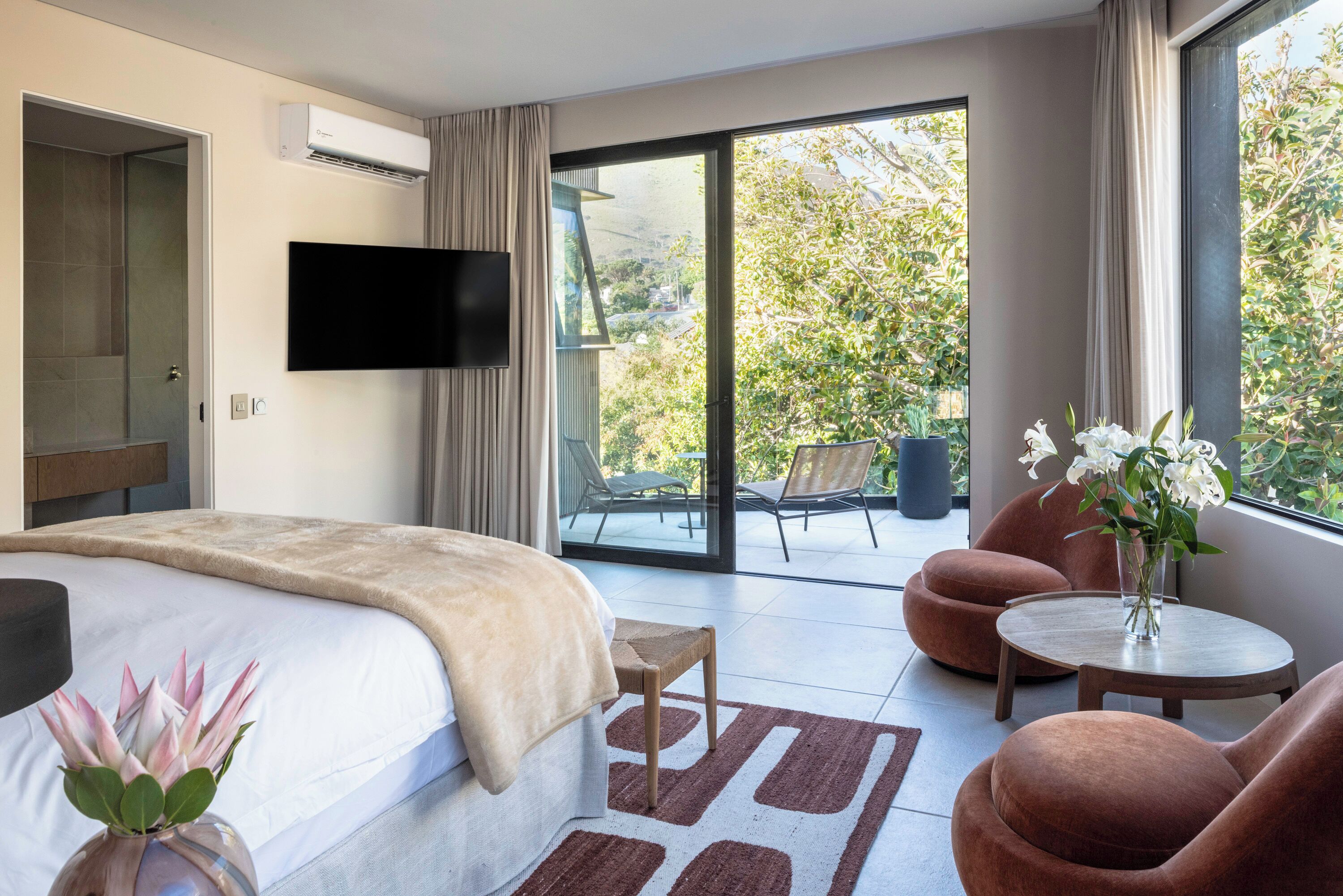 A modern hotel room with a large bed, a flat-screen TV, a balcony with chairs, and a view of greenery.