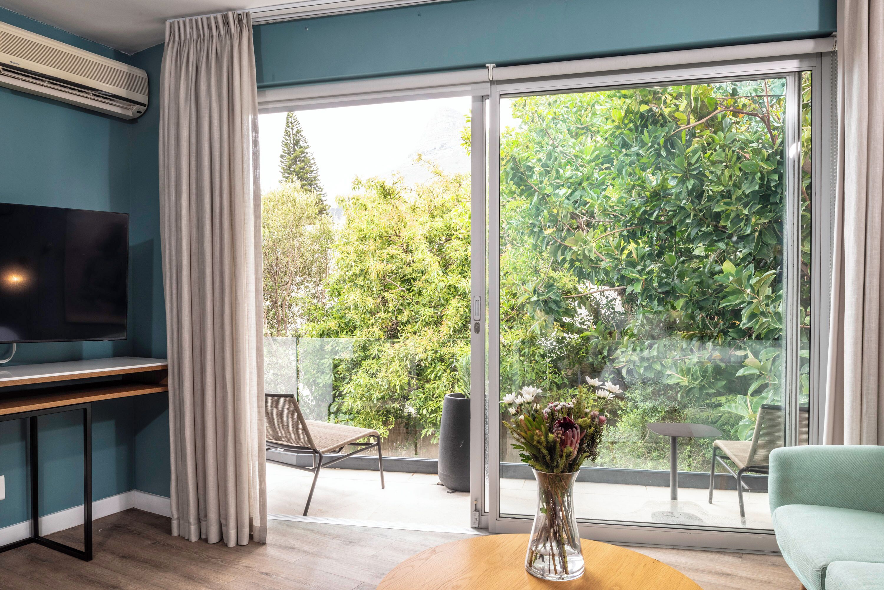Een moderne woonkamer met een flatscreen televisie, een houten tafel met een bloevas, een groene fauteuil en een grote schuifpui die leidt naar een balkon met tuinmeubilair.