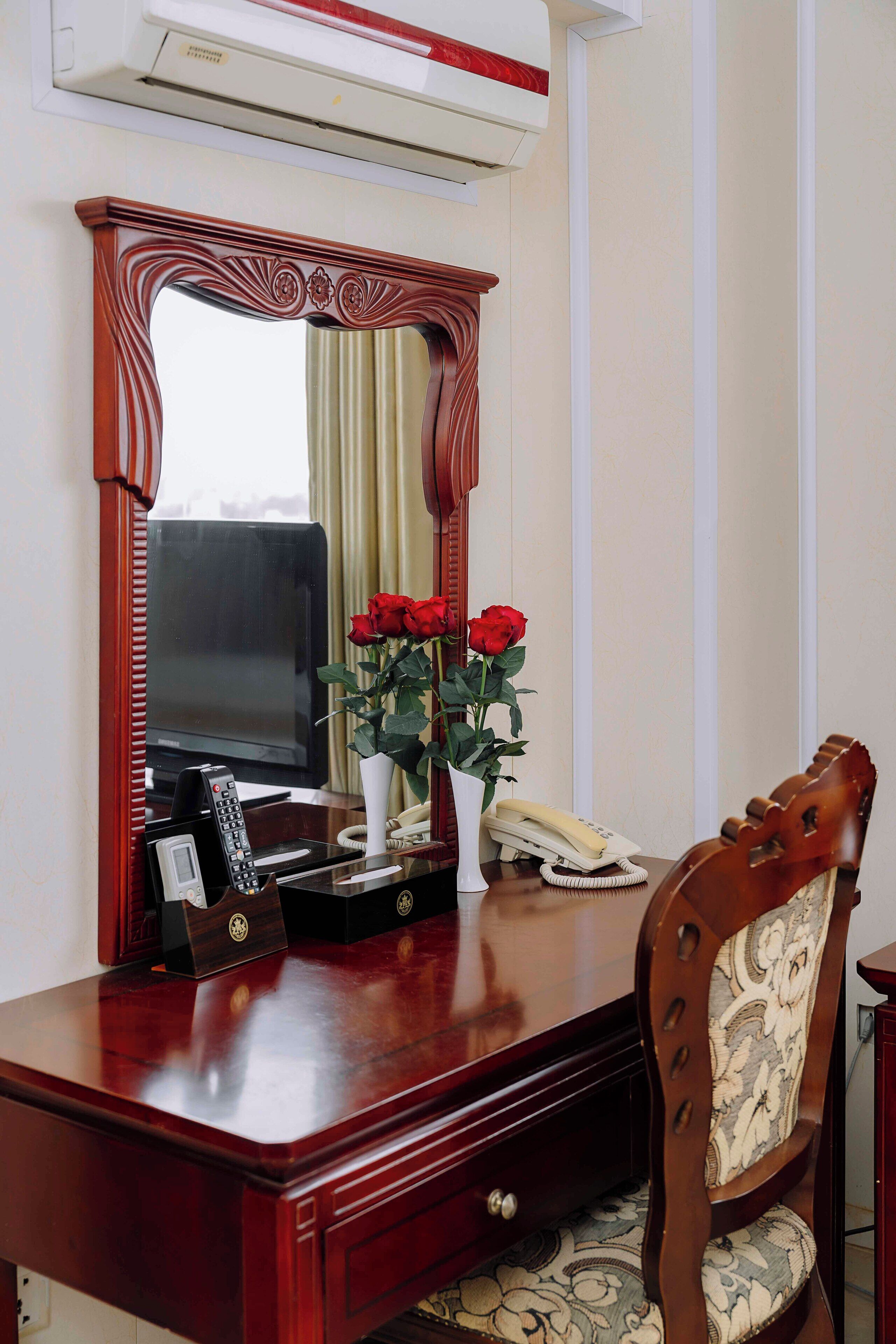 A wooden desk with a mirror, a vase with red roses, a telephone, and a television.