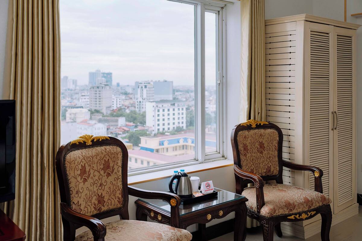 A hotel room with two ornate chairs, a small table with a coffee pot and cups, and a view of a cityscape through a large window.