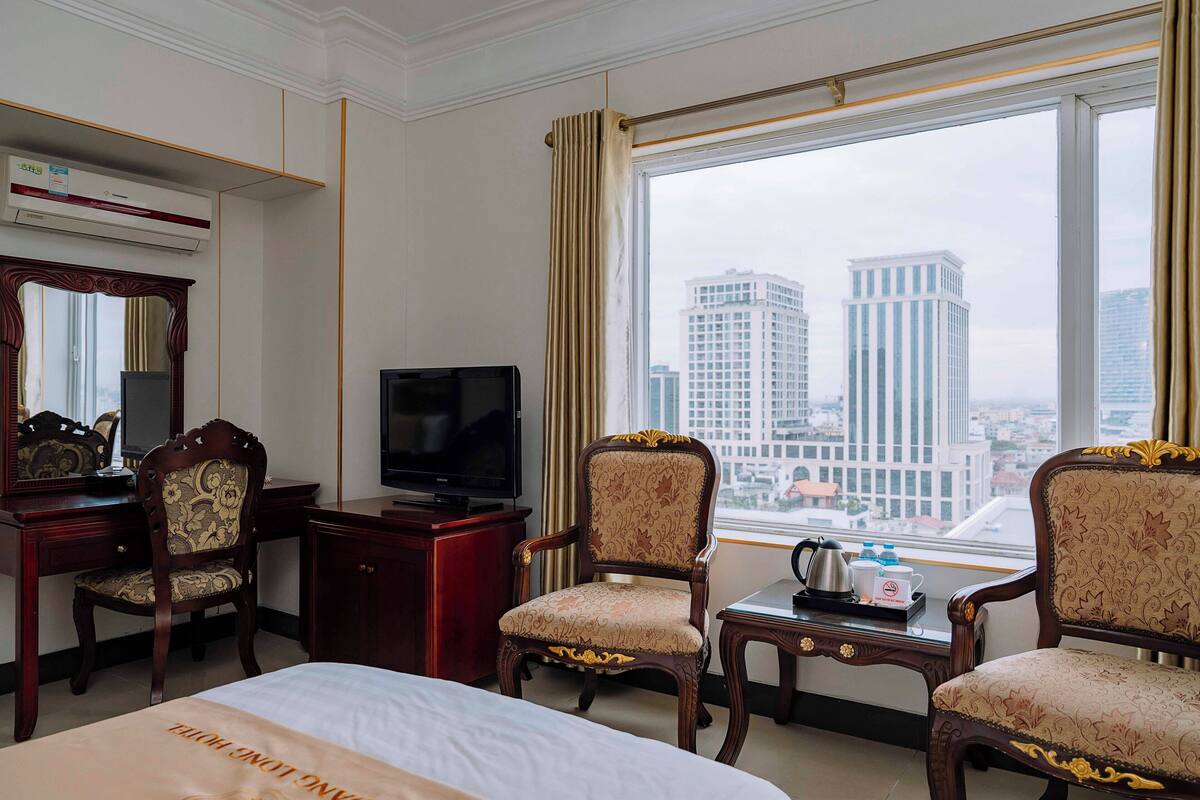 A hotel room with a bed, two armchairs, a desk with a television, and a window with a city view.