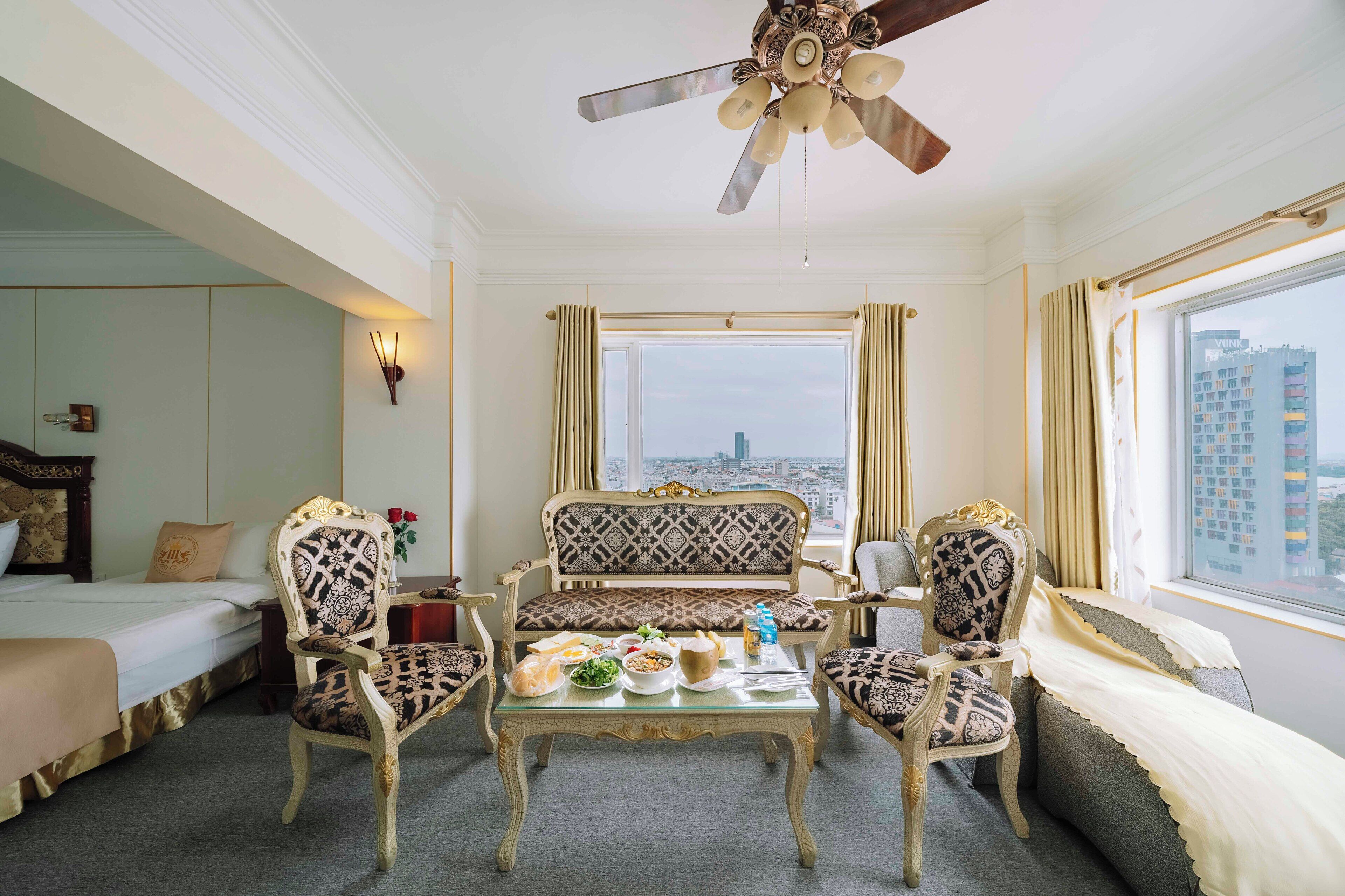 Ein Hotelzimmer mit einem Bett, einer Couch, zwei Sesseln, einem Esstisch mit Speisen, einem Deckenventilator und Blick auf eine Stadtlandschaft durch ein großes Fenster.