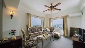 Quarto de hotel com uma janela grande, ventilador de teto, mesa de jantar com um vaso de flores e uma televisão em um armário de madeira.