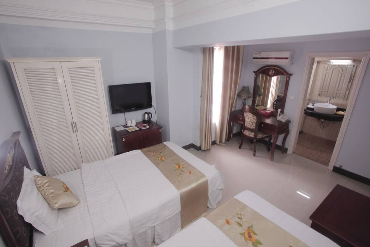 A hotel room with two beds, a television, a desk with a chair, a wardrobe, and a bathroom visible through an open door.