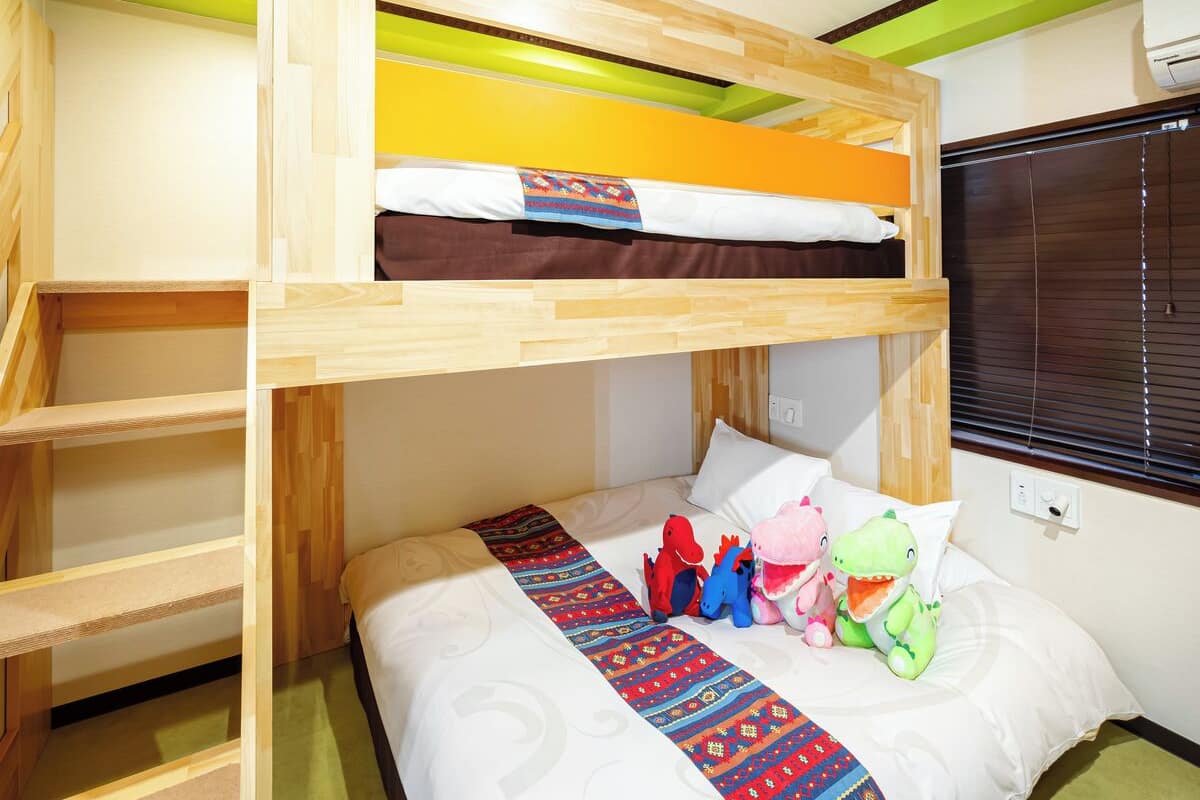 Een slaapkamer met een stapelbed, een houten ladder, een bed met een decoratieve spreideken en knuffeldiertjes op het onderste bed.