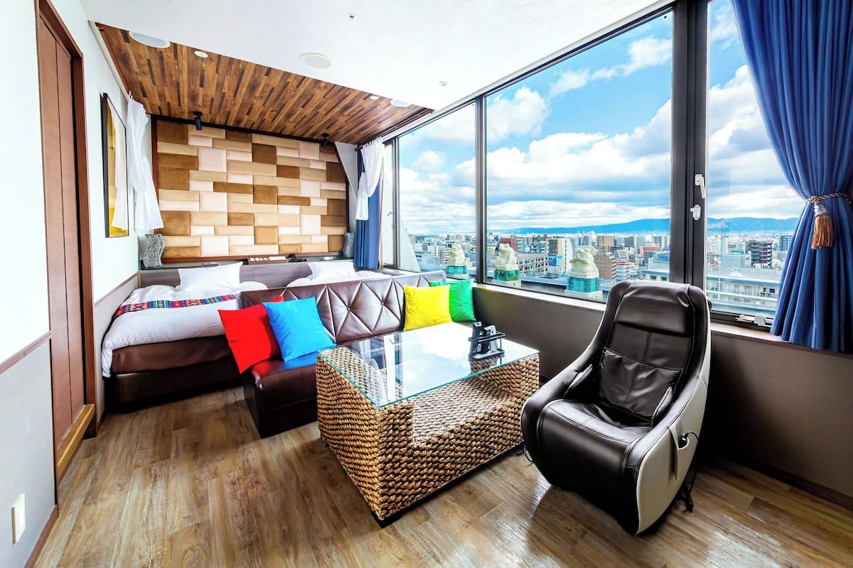 Een moderne hotelkamer met een bed, een glazen salontafel, een massagestoel en een groot raam met uitzicht op de stad.