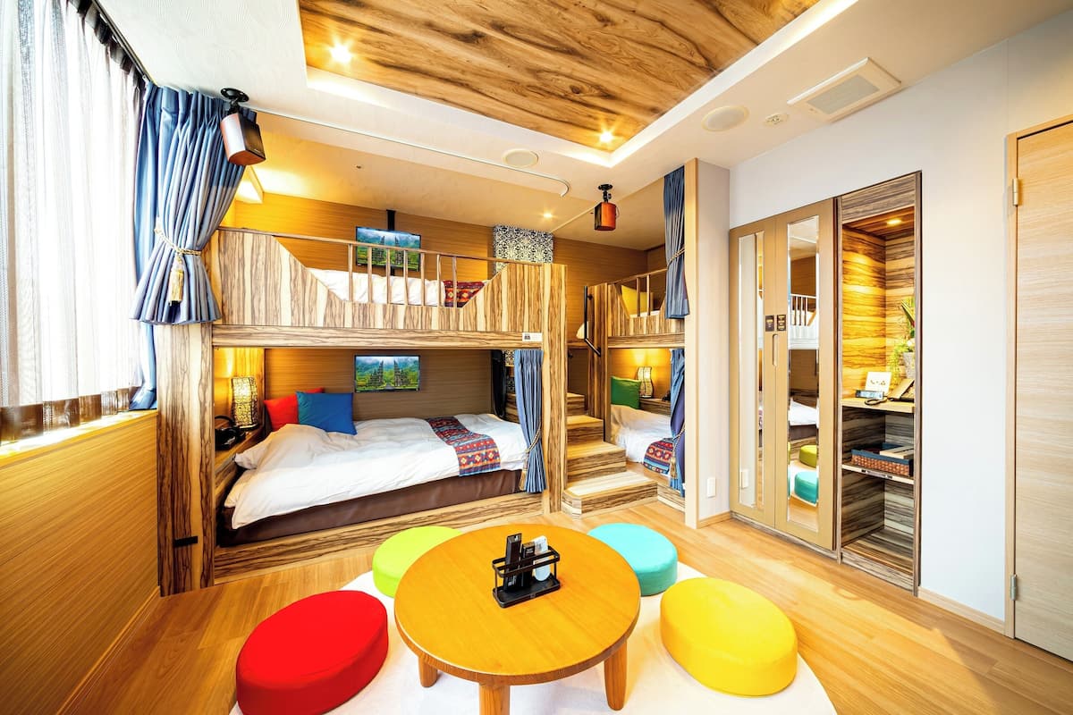 Een kleine kamer met stapelbedden, een ronde tafel en kleurrijke poefs.