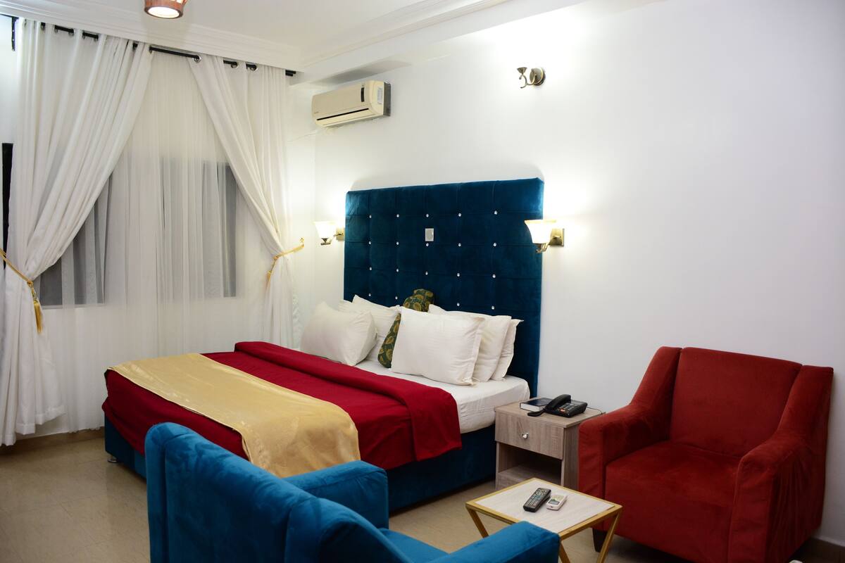 A hotel room with a bed, a red armchair, a blue armchair, a bedside table, and a nightstand.
