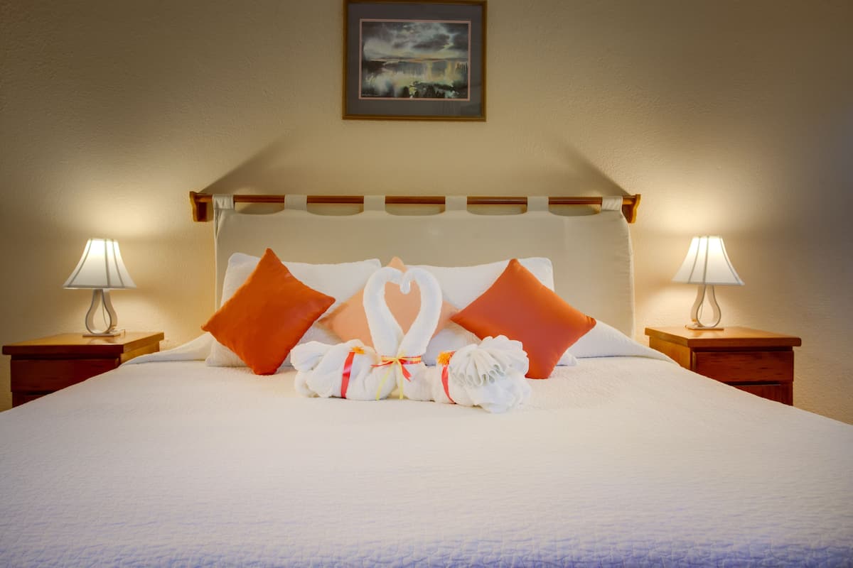 A neatly made bed with a heart-shaped towel arrangement and orange pillows, two bedside lamps, and a framed picture on the wall.