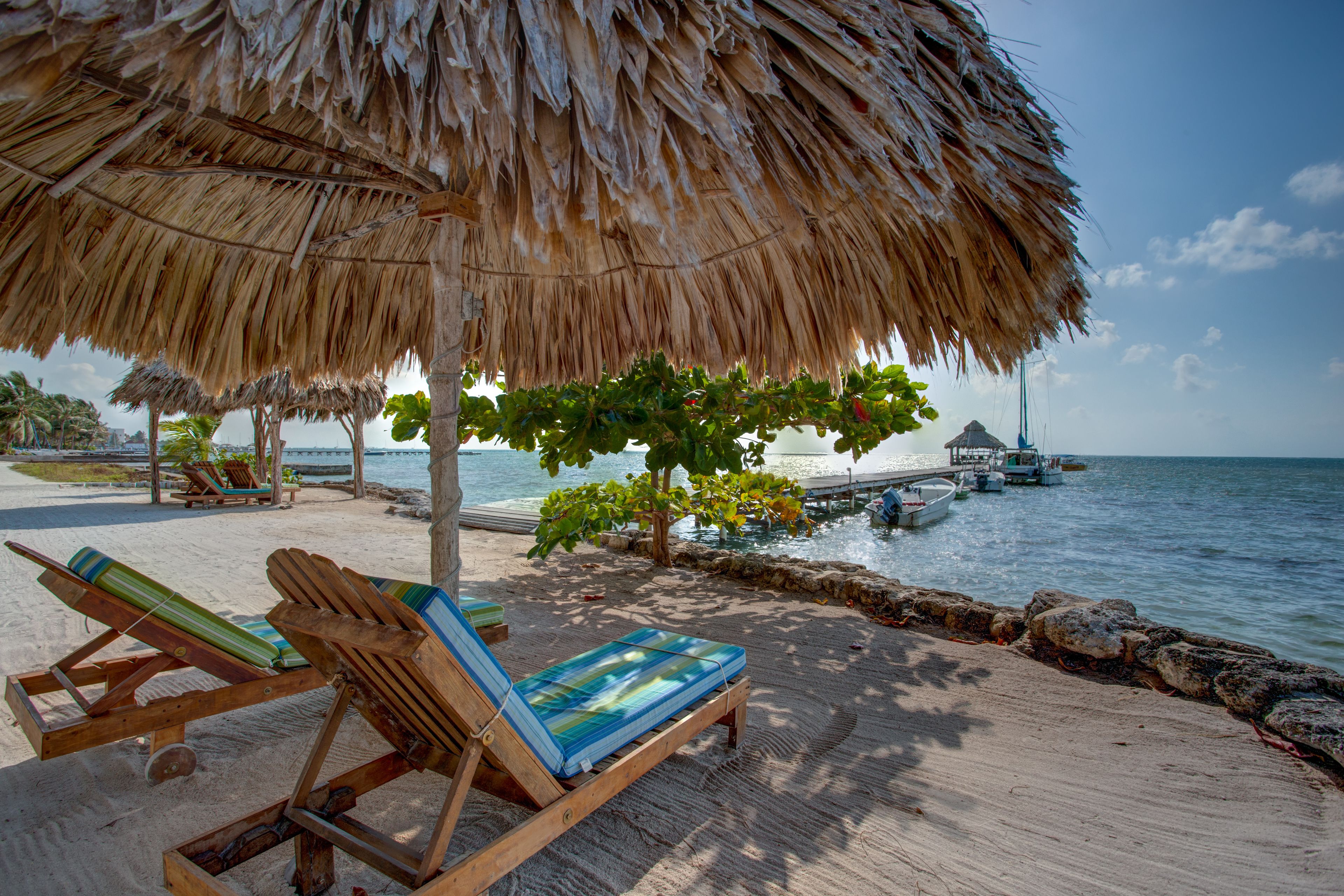 A beachside lounge with wooden chairs and a thatched umbrella, overlooking the sea and docked boats.