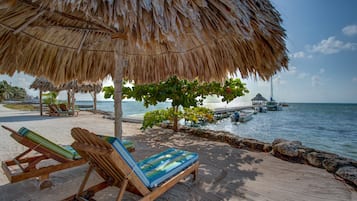 A beachside lounge with wooden chairs and a thatched umbrella, overlooking the sea and docked boats.