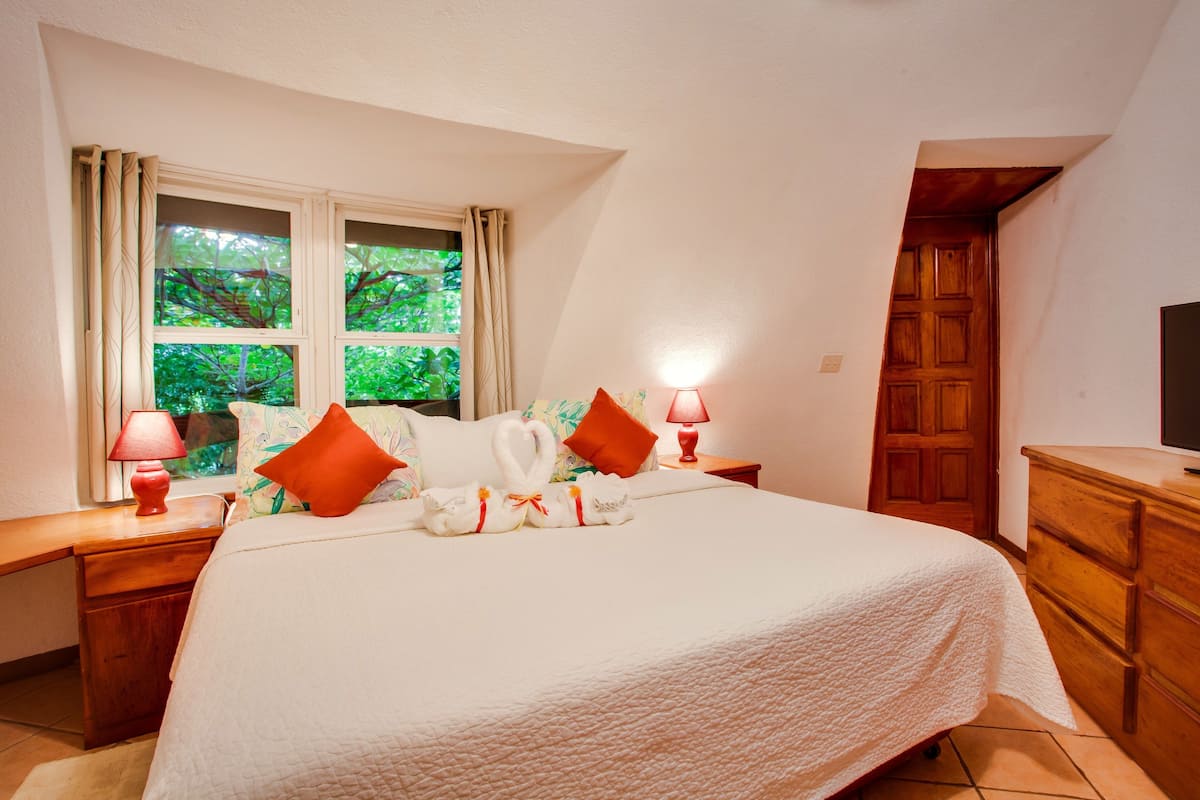 A neatly made bed with a white quilt, two red lamps on nightstands, a wooden dresser, and a window with curtains.