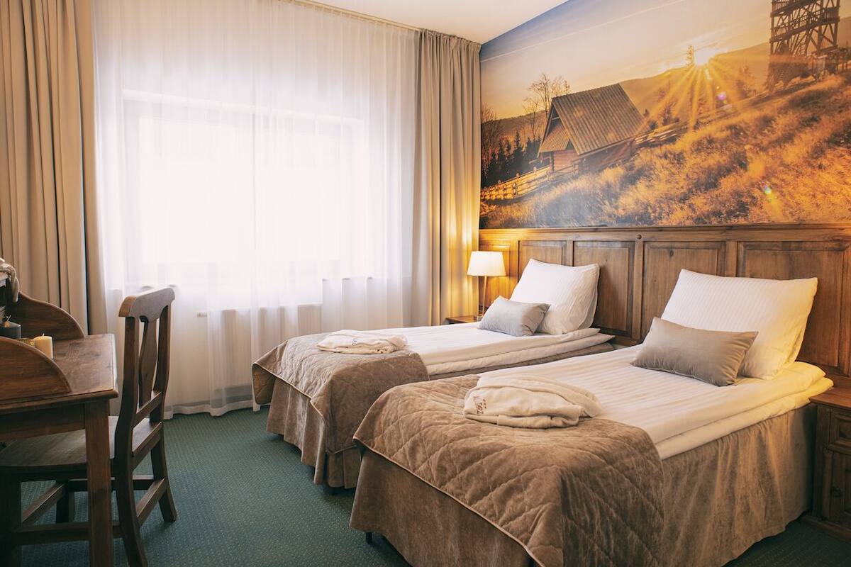 A hotel room with two beds, a desk, a chair, and a large window with sheer curtains.